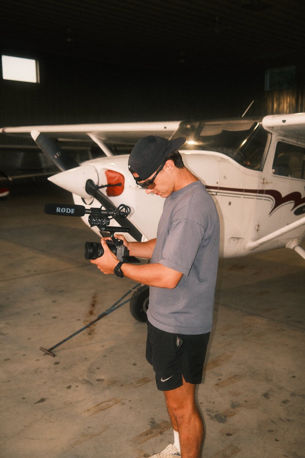 Gabriel en tournage, caméra à la main, devant un petit avion dans un hangar